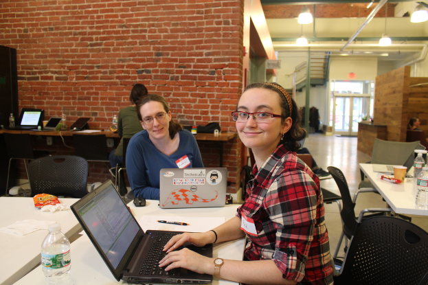 Zwei Frauen sitzen an einem Tisch mit Laptops, einer Flasche, einem Stift und Papier in einem Büro mit einer roten Backsteinwand, Deckenleuchten, einer Treppe und einer Eingangstür im Hintergrund.
