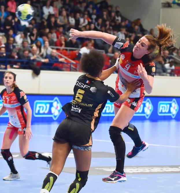 Eine Gruppe von Frauen, die auf einem Feld Handball spielen, während Zuschauer von Stühlen, Geländern und einer Wand im Hintergrund zuschauen.