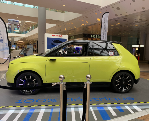 Gelber wasserstoffbetriebenes Elektroauto vor einem Gebäude mit Absperrpollern, Seilen, Bannern, einer Person im Auto, einer Säule, einer Gruppe von stehenden Menschen, Schildern, einer Topfpflanze, Deckenleuchten und Dachventilatoren.