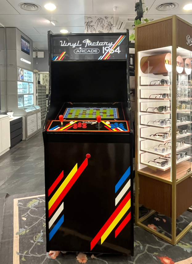 Ein Geschäft mit einem zentralen Retro-Arcade-Automaten, einem Regal mit Gläsern auf der rechten Seite und Hintergrundelementen wie Schränken, einem Fernseher und anderen Gegenständen, mit Deckenlampen, die eine Retro-Atmosphäre schaffen.