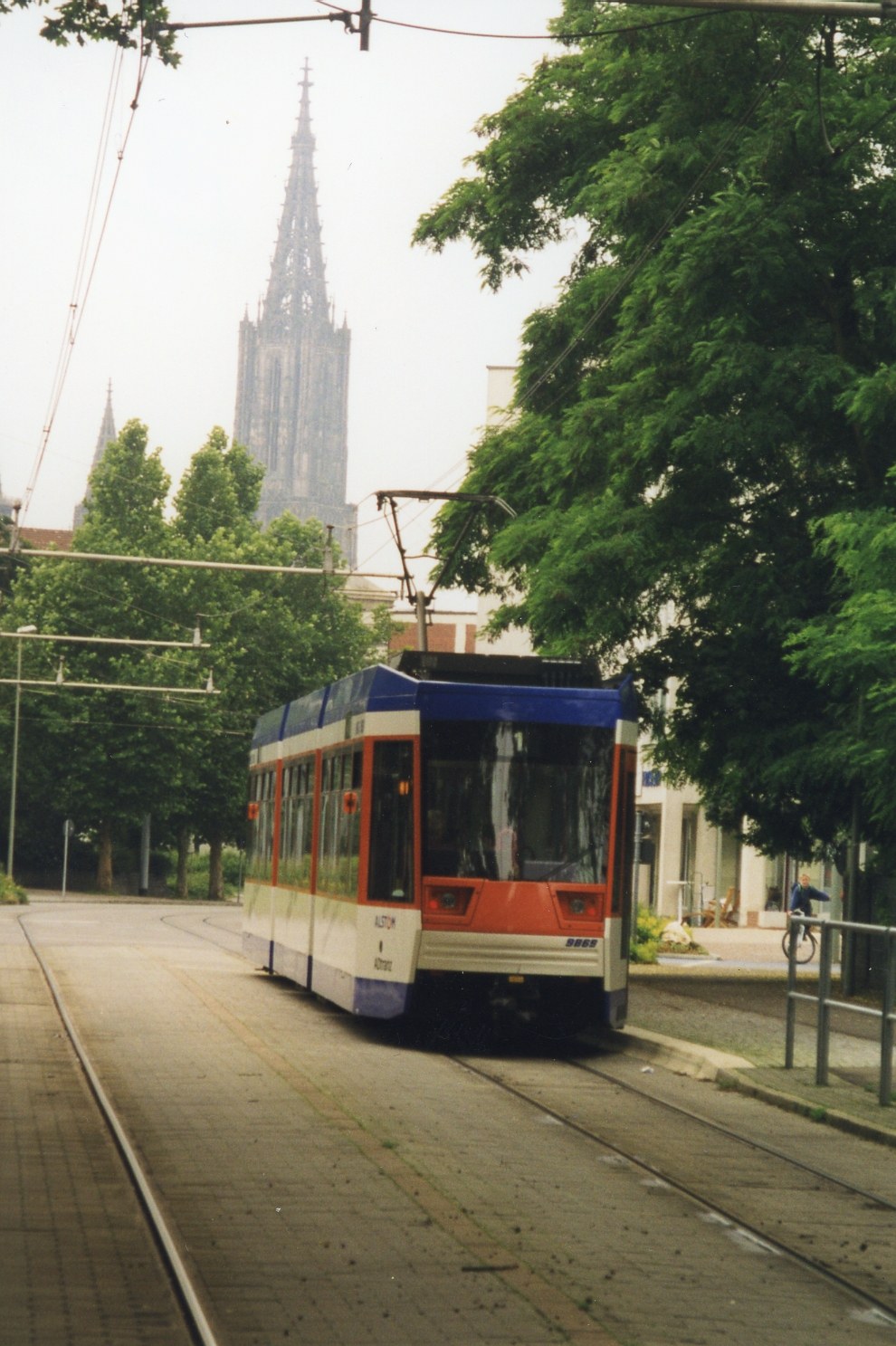 Rotes und weißes Zugfahrzeug auf Schienen neben einem hohen Gebäude, mit einer Person, die ein Fahrrad auf dem benachbarten Gehweg fährt, umgeben von Bäumen und Gebäuden unter einem klaren blauen Himmel.