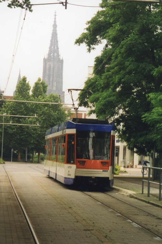 Rotes und weißes Zugfahrzeug auf Schienen neben einem hohen Gebäude, mit einer Person, die ein Fahrrad auf dem benachbarten Gehweg fährt, umgeben von Bäumen und Gebäuden unter einem klaren blauen Himmel.