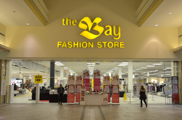 Außenansicht eines Bay Fashion Stores in einem Einkaufszentrum mit Menschen, Texttafeln an Wänden, Säulen, Deckenleuchten und Hintergrundanzeigen.