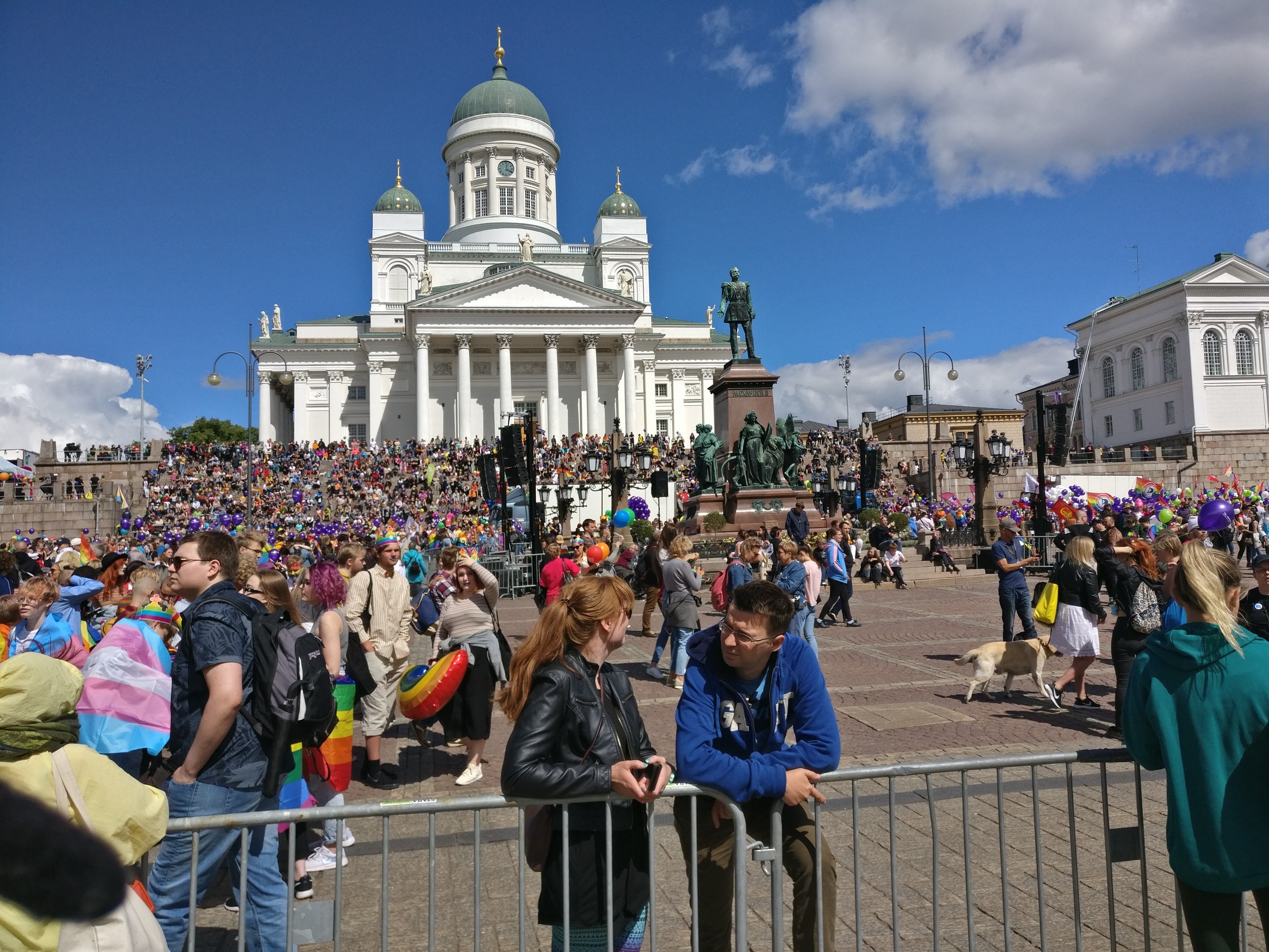 A large crowd stands outside a building with windows, pillars, arches, railings, light poles, and statues, surrounded by trees under a cloudy sky, with some people carrying bags and balloons, indicating a gay pride parade in Helsinki.
