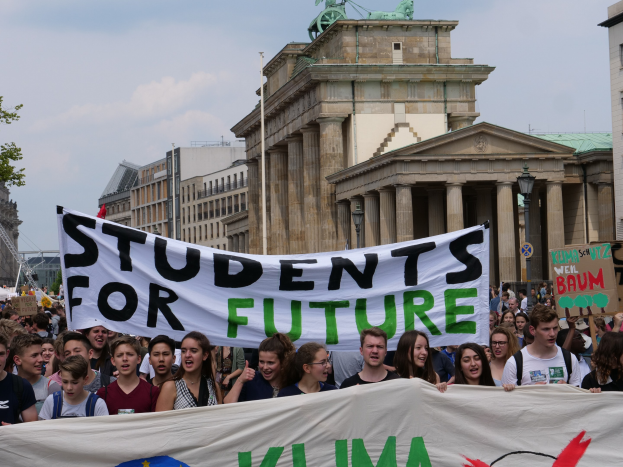 Gruppe von Schülern marschiert in Berlin mit einem leuchtend bunten "Students for Future"-Schild gegen Gebäude, Bäume und Himmel.