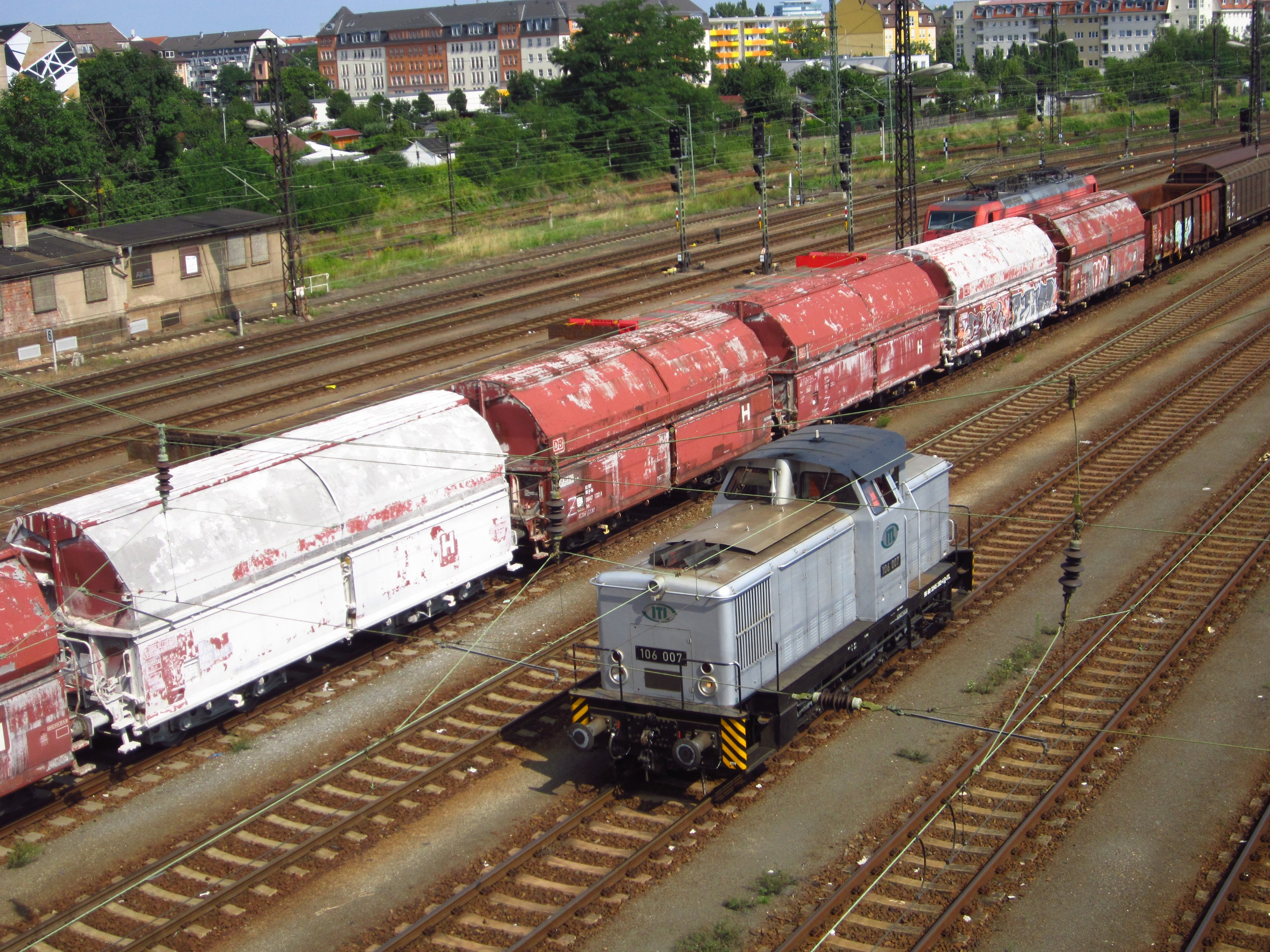 Freight train traveling on tracks beside a city with electric poles and cables, buildings, trees, and a clear blue sky in the background.