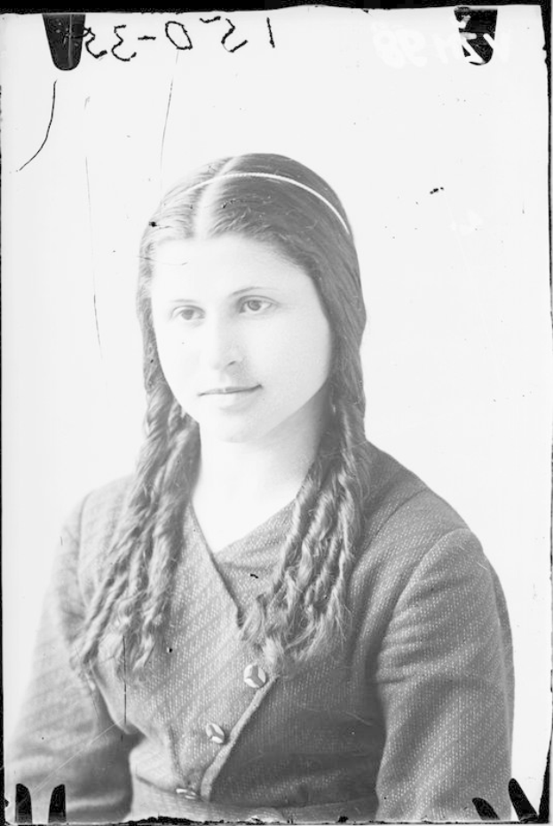 Ein Schwarz-Weiß-Foto einer Frau mit langen Zöpfen, identifiziert als Julia Margaret Cameron, die ein weißes Kleid mit einem gemusterten Schultertuch trägt, ihr Haar ist zurückgebunden und sie hat einen sanften Gesichtsausdruck und ihre Hände sind vor der Brust verschränkt.