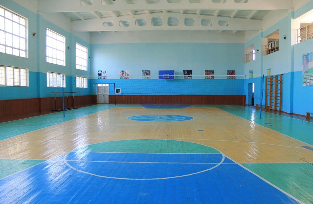 Großer Turnsaal mit blau-weißem Boden, der einen Basketballplatz, Netz, Stangen, eine Tür, Fenster, Plakate an den Wänden, ein Geländer und ein Dach enthält.