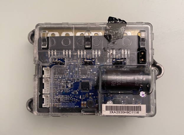 Close-up of a small electronic circuit board with an attached battery housed in a plastic case on a white surface.