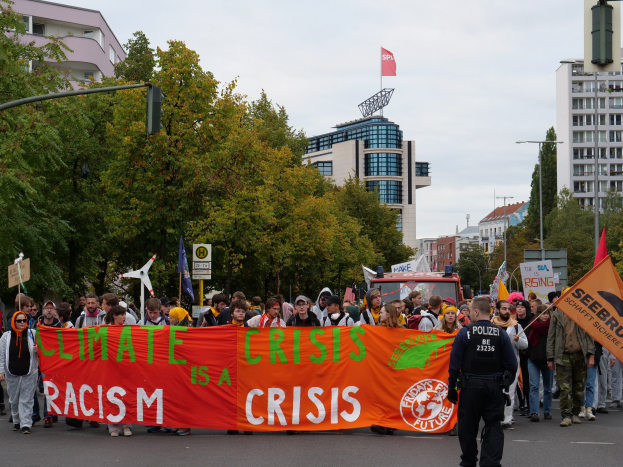 Eine Gruppe von Menschen marschiert eine baumbestandene Straße entlang und hält ein Transparent mit der Aufschrift "Klima-Krise ist eine Krise", mit Gebäuden und einem klaren blauen Himmel im Hintergrund.