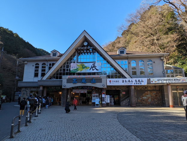 Eine Gruppe von Menschen steht ├Ąu├čer einem Geb├Ąude mit Fenstern, Schildern, Bannern, einer Uhr, Metallstangen, einem Zaun, einem Stra├čenpfahl, B├Ąumen, H├Ąngen und einem bew├Âlktem Himmel, einige tragen Taschen in der N├Ąhe von beliebten Touristenattraktionen.