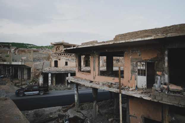 Ein Lastwagen fährt auf einer Straße neben einem zerbombten Gebäude mit zerbrochenen Wänden und verstreuter Trömmern, mit anderen Gebäuden, Masten, Bäumen, einem Hügel und dem Himmel im Hintergrund.