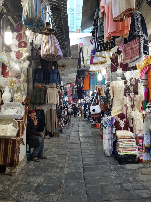 A bustling street market in Jerusalem's old city with stalls selling clothes, bags, lights, and other items, as people browse and interact in the background.