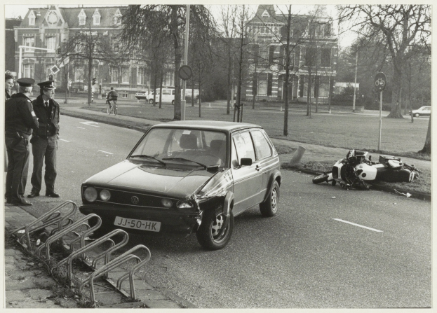 Ein Schwarz-Weiß-Foto eines Autos, das auf der Seite der Straße in ein Motorrad gekracht ist, mit drei Personen auf dem Gehweg links und Bäumen, Pfählen, Gebäuden und anderen Fahrzeugen im Hintergrund.