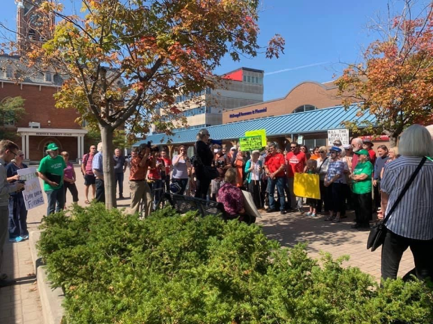 Gruppe von Studentinnen und Studenten protestiert vor einem Gebäude der University of Michigan, hält Schilder und Kameras hoch, mit Bäumen und Himmel im Hintergrund.