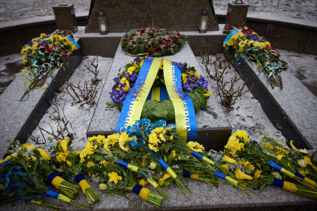 Gedenkstätte in Kiew, Ukraine mit einem Blumenkranz auf einem Grab, das mit einer blauen Schleife markiert ist und von zusätzlichen Blumensträußen umgeben ist, vor einem schneebedeckten Hintergrund.