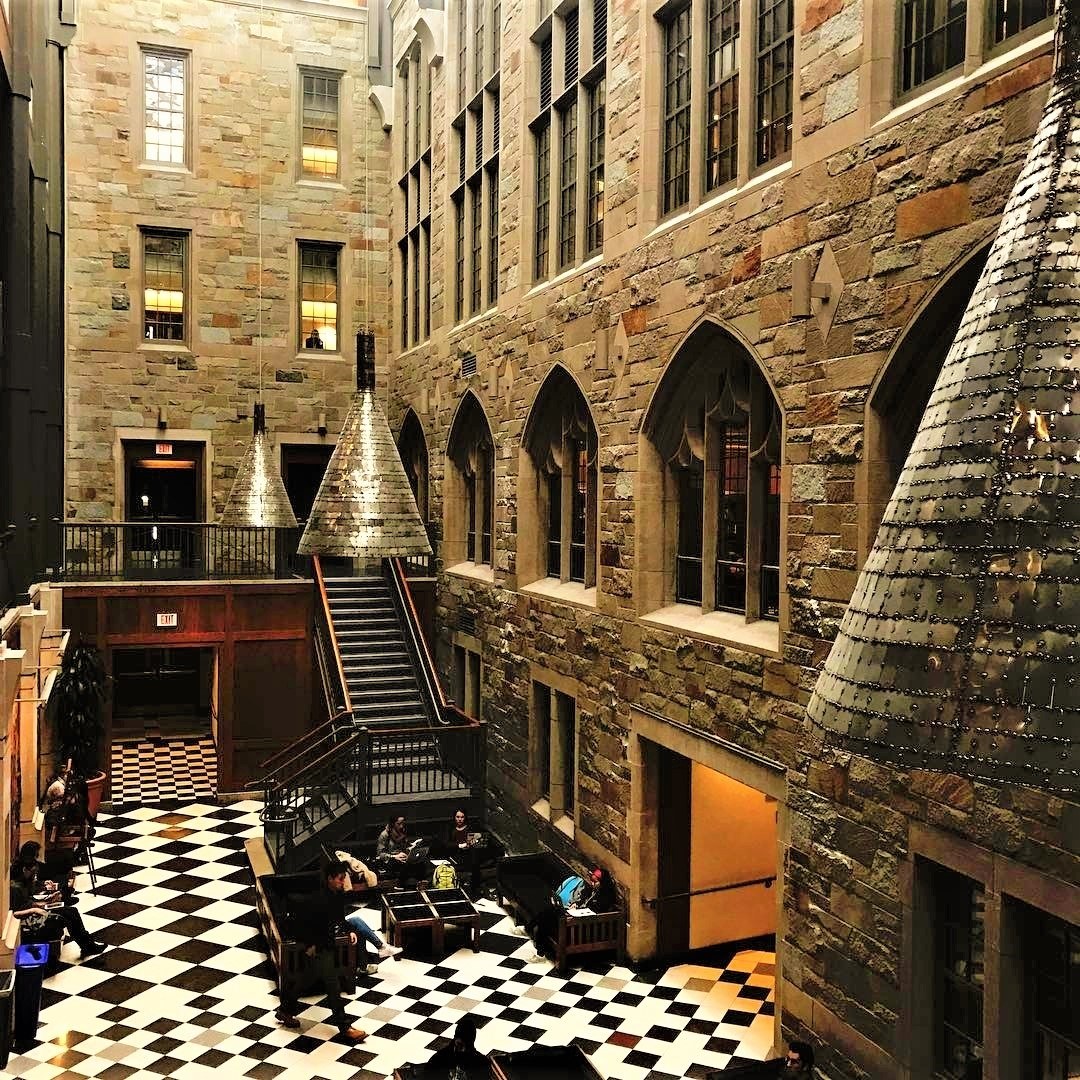 Large room with black and white checkered floor, central staircase with railings, windows, overhead lights, and several people seated at tables and chairs, resembling a Harry Potter studio tour in London.