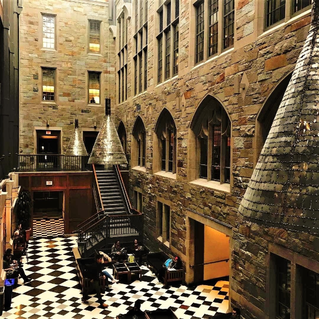 Large room with black and white checkered floor, central staircase with railings, windows, overhead lights, and several people seated at tables and chairs, resembling a Harry Potter studio tour in London.