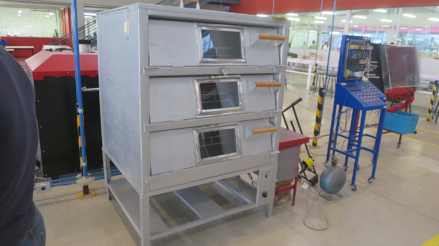 Ein Labor mit elektrischen Geräten und Metallregalen im Vordergrund, eine Person links und eine Glaswand mit ein paar Personen dahinter rechts.