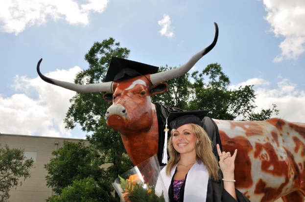 Eine Frau in einer Graduationskappe und -robe lächelt und hält einen Strauß Blumen, mit einer Skulptur, Bäumen, einem Gebäude und einem bewölkten Himmel im Hintergrund.