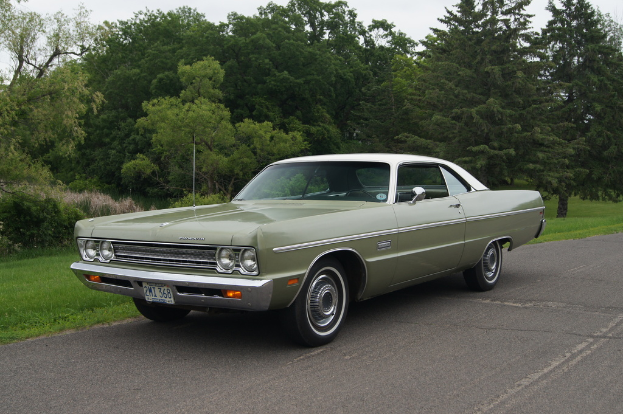 Ein grünes Oldtimer-Auto fährt auf einer Straße mit Bäumen und Wiesen im Hintergrund unter einem klaren Himmel.