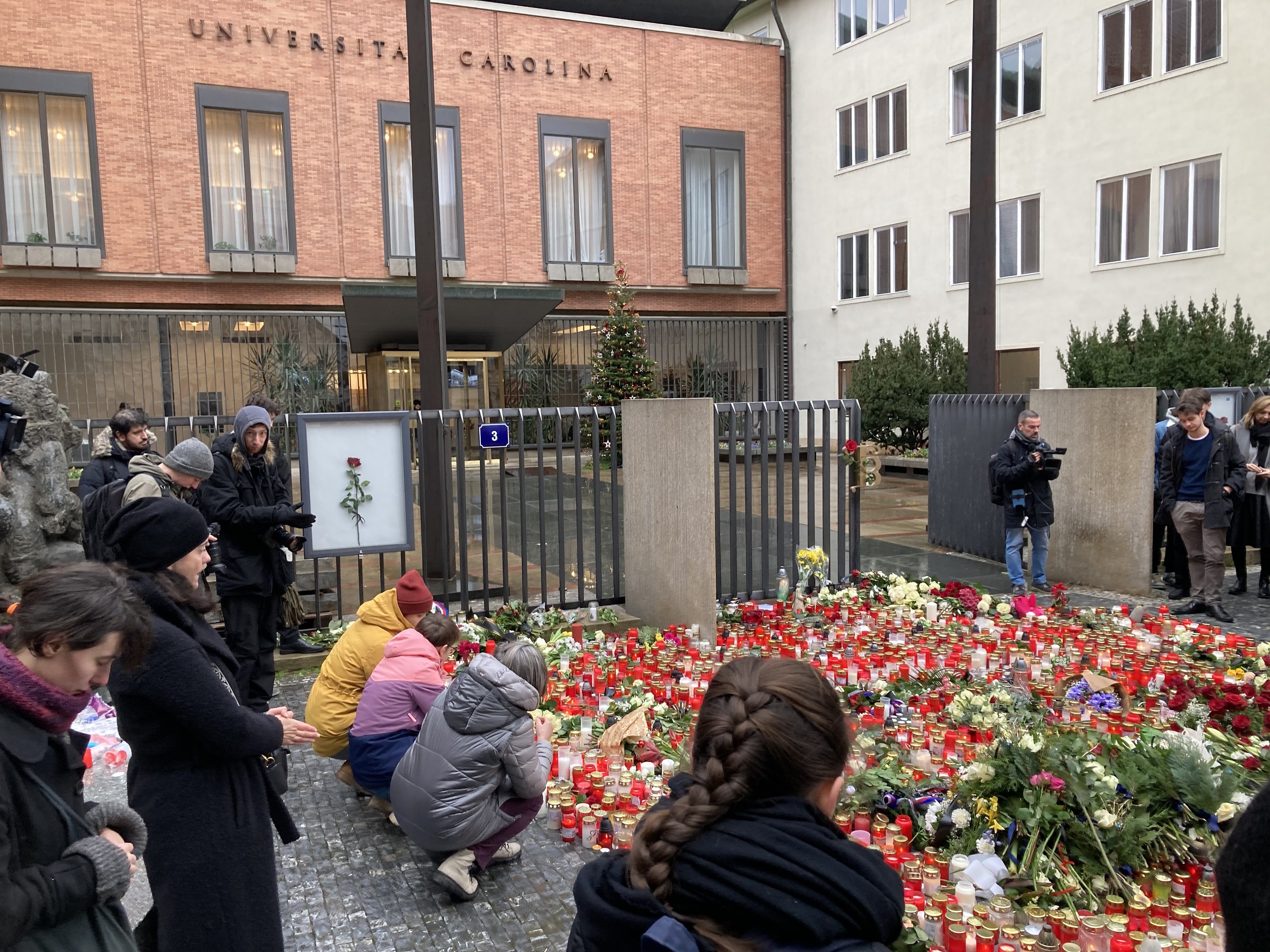 A group of people stands around flowers and candles placed on the ground outside a building, some wearing caps and holding cameras or bouquets, with buildings, trees, and railings in the background.