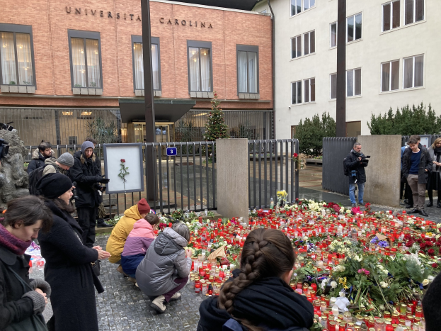 A group of people stands around flowers and candles placed on the ground outside a building, some wearing caps and holding cameras or bouquets, with buildings, trees, and railings in the background.