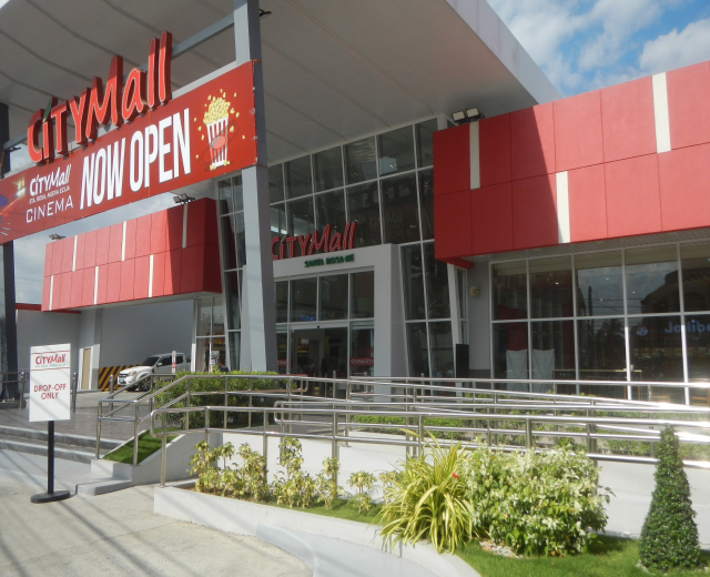 Eingang eines Stadtzentrums mit einem "Stadtzentrum jetzt geöffnet"-Schild, umgeben von Grün, einem Metallzaun, Menschen, einem Auto, Pfosten, einer Fahne, Lichtern und einem bewölkten Himmel.