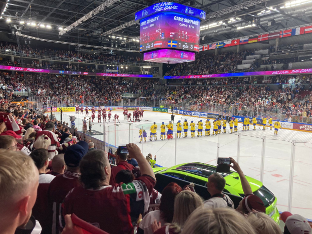 Ein Eishockeyspiel in einer großen Arena mit Spielern auf dem Eis, Zuschauern an den Seiten, Fotografen mit Kameras, Arena-Beleuchtung und einem Bildschirm mit Text über dem Eis.