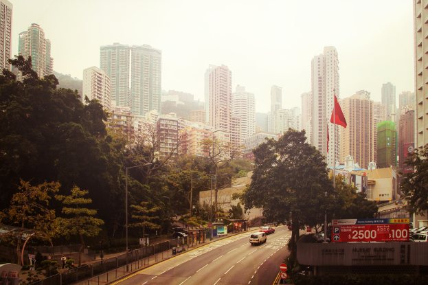 Fahrzeuge auf einer Straße mit Pfosten, Fahnen und einer Werbetafel auf der rechten Seite und Bäumen, Gebäuden, Hochhäusern und Himmel auf der linken Seite.