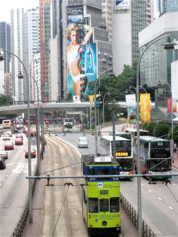 Eine belebte Stadtstraße mit verschiedenen Fahrzeugen, darunter Doppeldeckerbusse, Autos und andere Fahrzeuge, umgeben von Pfählen, Lampen, Tafeln, Bannern, Bäumen, einer Brücke, hohen Gebäuden und einem klaren blauen Himmel, mit Text am unteren Bildrand.