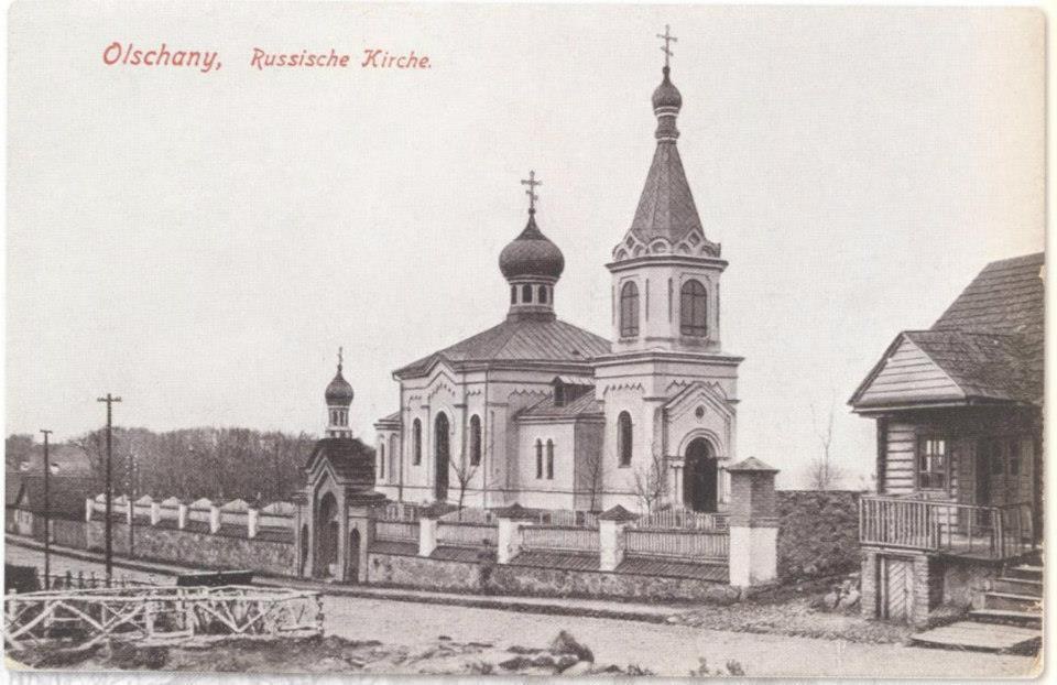 Schwarzes und weißes Foto einer russisch-orthodoxen Kirche in Olschany, Russland, umgeben von Häusern, Bäumen, Pfählen und einem Zaun unter einem klaren Himmel, mit Text oben.