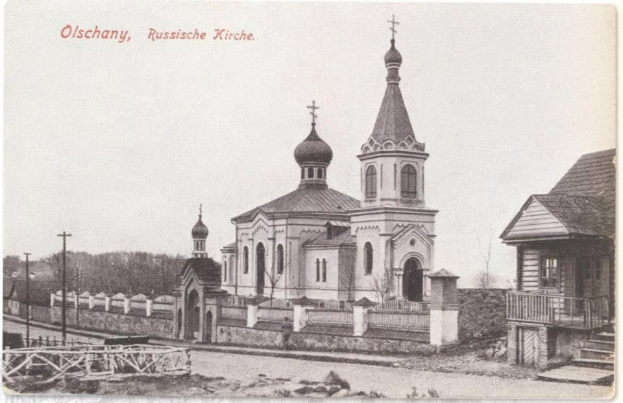 Schwarzes und weißes Foto einer russisch-orthodoxen Kirche in Olschany, Russland, umgeben von Häusern, Bäumen, Pfählen und einem Zaun unter einem klaren Himmel, mit Text oben.