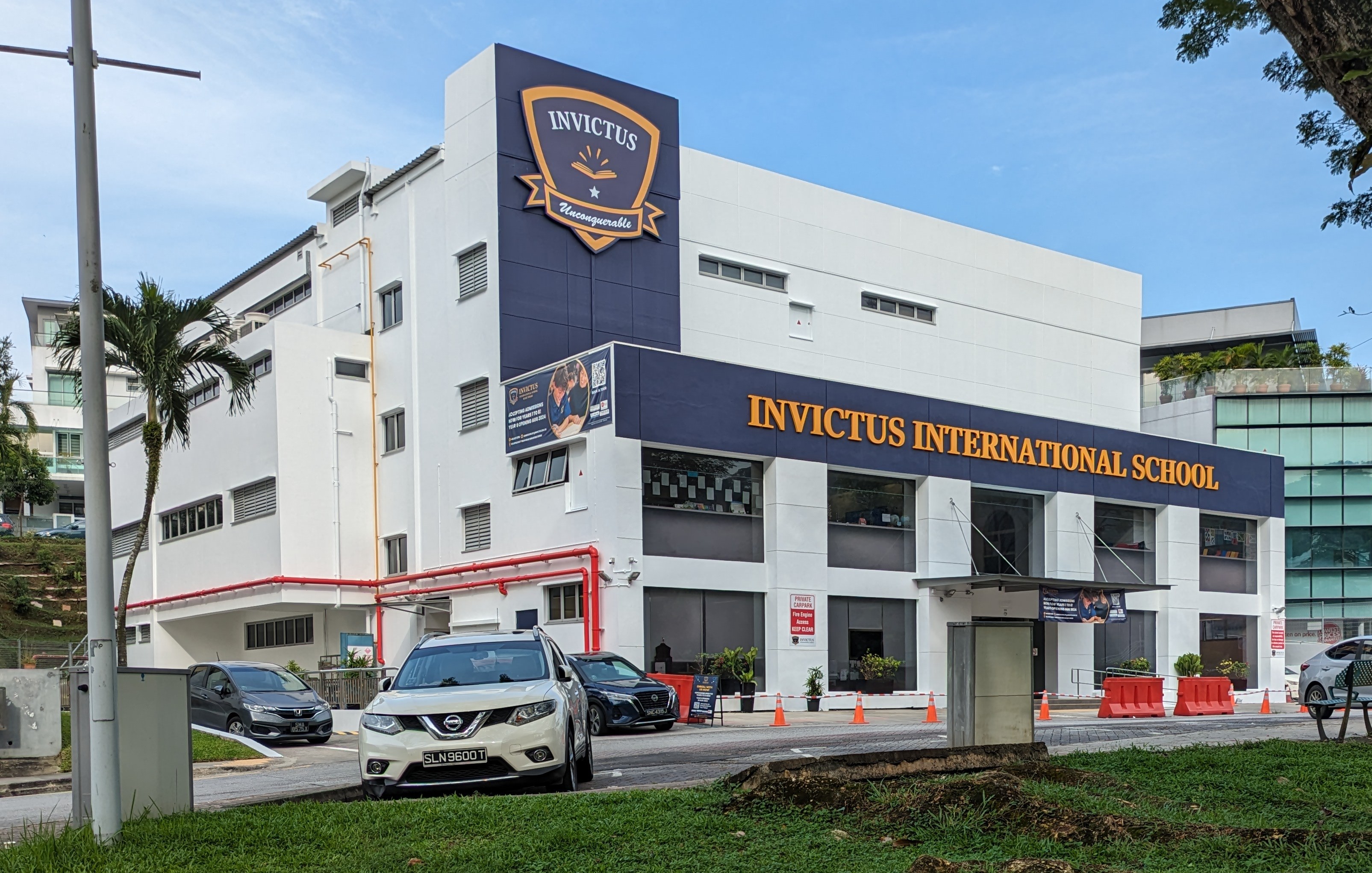 Exterior view of Invictus International School in Singapore, featuring buildings, signage, vehicles, road infrastructure, greenery, and a cloudy sky.