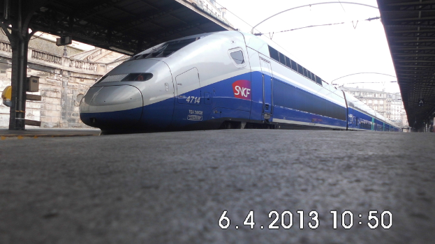 Ein Zug ist in der Mitte des Bildes zu sehen, mit einem Bahnsteig und Text unten, Gebäuden und einem Dach links und Gebäuden, Dächern, Kabeln und Himmel oben.