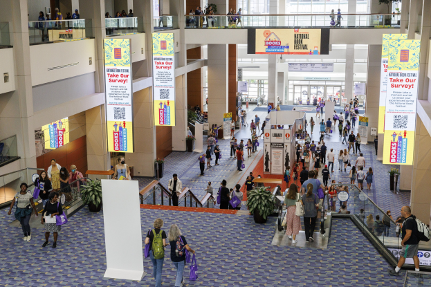 Eine große Menschenmenge geht durch ein großes Gebäude mit Bannern, Topfpflanzen, Rolltreppen, Geländern, Säulen, bedruckten Tafeln, Glastüren und anderen Gegenständen, einige tragen Taschen, was auf den Besuch der Nationalen Buchmesse hinweist.