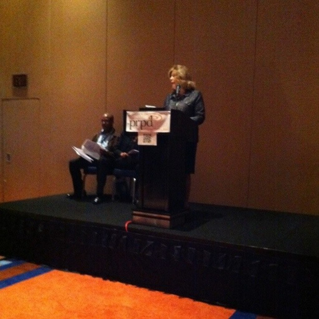 Zwei Männer sitzen auf Stühlen, während eine Frau vor einem Rednerpult auf einer Bühne steht, mit einer hölzernen Wand im Hintergrund und einem Teppich im Vordergrund.