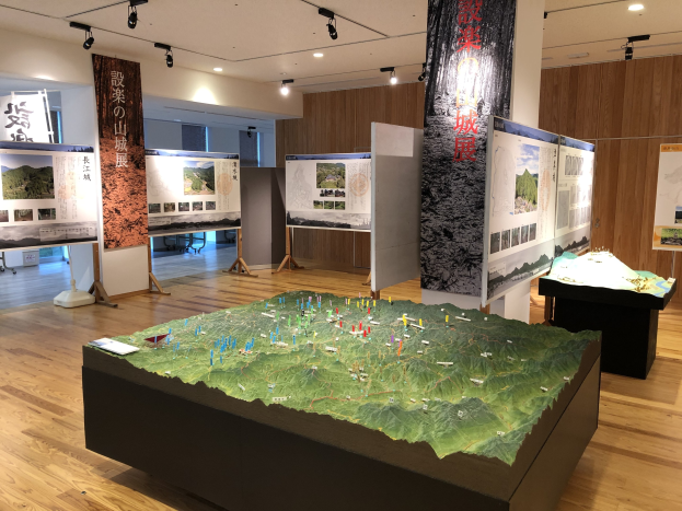 Ausstellungsraum mit verschiedenen historischen Karten Japans, darunter aufgehängte Banner, Tischausstellungen, beleuchtete Tafeln und eine hölzerne Rückwand.