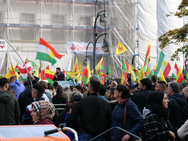 Eine große Gruppe von Menschen mit Fahnen und einer Person mit einem Mikrofon vor einem Gebäude, mit einem Fahrzeug, Baum, Pfosten, Lampen und Schildern mit Text im Hintergrund, wahrscheinlich bei einer kurdischen Demonstration in Istanbul.