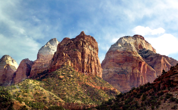 Ein malerischer Blick auf den Zion-Nationalpark in Utah mit majestätischen Bergen, grünen Bäumen, steinigem Gelände und einem Himmel mit weißen, flauschigen Wolken.