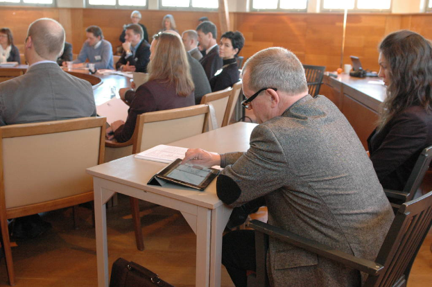 Zwei Personen sitzen auf schwarzen Stühlen an einem runden Tisch in einem Bürokongressraum, ein Mann nutzt ein Tablet und eine Frau hält ein Buch, mit anderen Personen, ID-Karten, Gläsern, Wasser und einem Telefon im Bild.