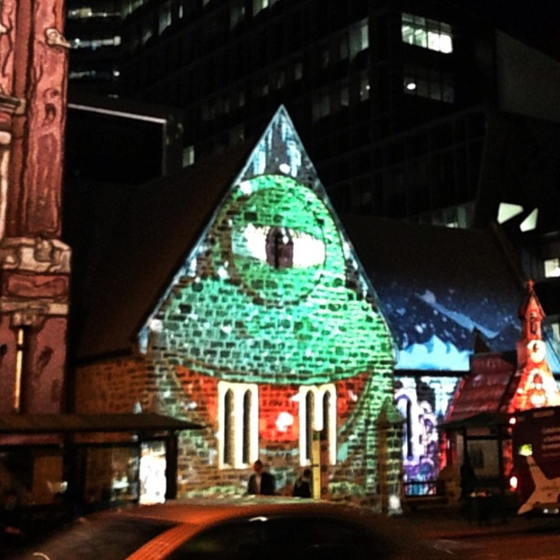 Ein zentrales Haus mit einer Person in der Nähe, flankiert von Häusern auf beiden Seiten, ein Gebäude mit Lichtern im Hintergrund und ein Auto unten.