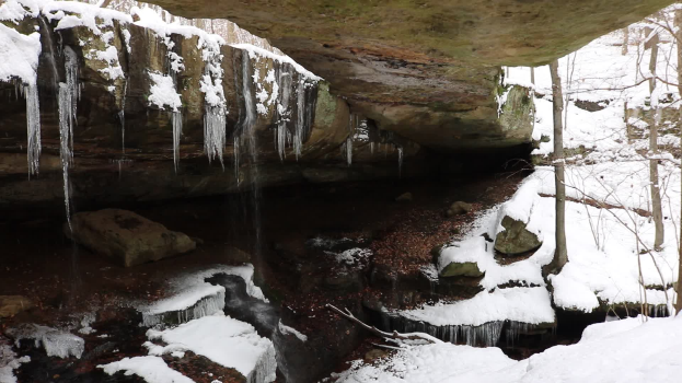 Ein kleiner Wasserfall ergießt sich über einen schneebedeckten, steinigen Hang in einem bewaldeten Gebiet, mit Eiszapfen an den Felsen und schneebedeckten Bäumen im Hintergrund.