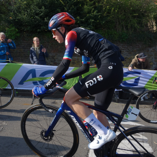 Profi-Radsportlerin Katie Archibald mit Helm, Schutzbrille, Handschuhen und Schuhen auf einem Fahrrad die Straße entlang neben einer Menschenmenge fahrend, mit einem Banner, einer Wand, Bäumen und einem Gebäude im Hintergrund.