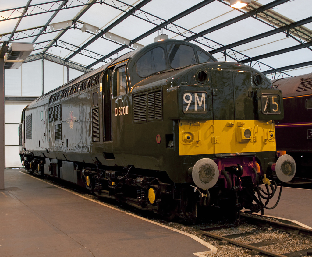 Ein Zug fährt auf den Schienen neben einem Bahnsteig mit einer Stange auf dem Dach, die Metallstangen mit Lampen trägt.