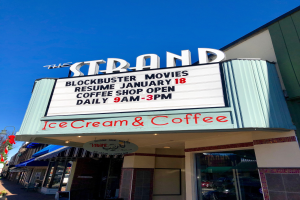 Der Strand-Blockbuster-Movies-Start am 18. Januar Kaffee-Shop-Gebäude mit einem Namensschild, einem Straßenschild, einer Gruppe von Bäumen, einigen Topfpflanzen und einem bewölkten Himmel.