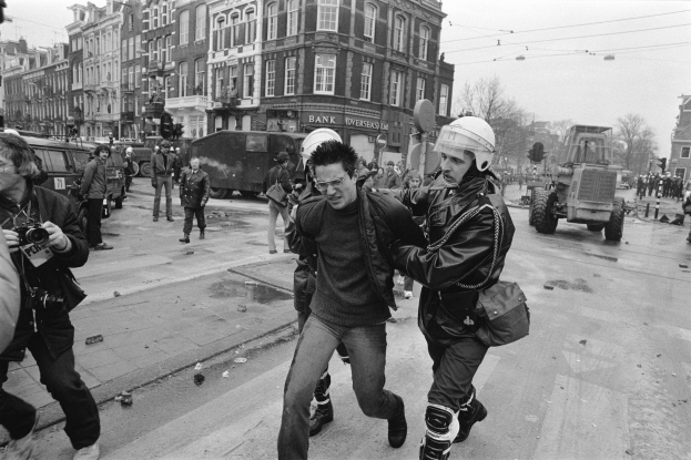 Ein Schwarz-Weiß-Bild eines Mannes, der von einem Polizeibeamten auf einer Straße eskortiert wird, mit einer Gruppe von Menschen mit Kameras, Fahrzeugen, Schildern, Straßenlaternen, Ampeln, Kabeln, Bäumen, Gebäuden mit Fenstern und einer bewölkten Himmel.