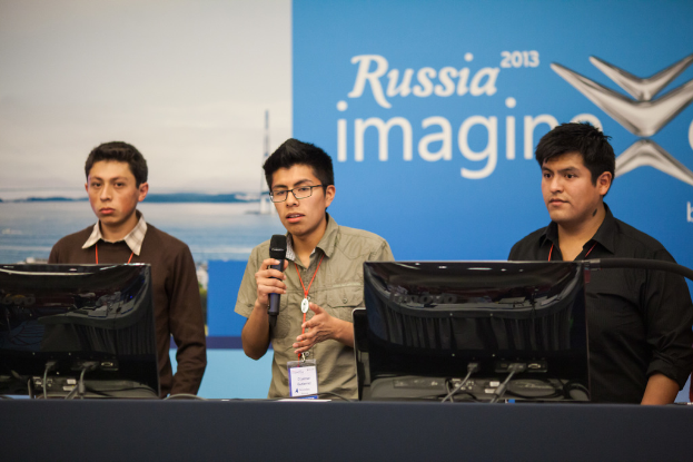 Drei Jungs stehen neben einem Schreibtisch mit zwei Computersystemen, einer spricht in ein Mikrofon und trägt ID-Karten mit roten Tags, vor einer Wand mit einer Werbetafel, auf der "Russia Imagine 2013" steht.