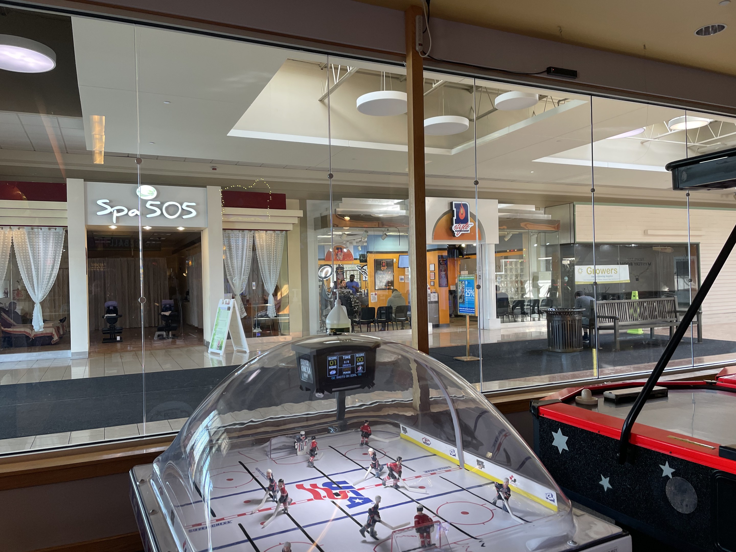 Eine Eishalle in einem Einkaufszentrum, umgeben von Glaswänden mit Menschen, Möbeln und Auslagen, die durch die Glaswände sichtbar sind, und einem Air-Hockey-Tisch auf der rechten Seite.