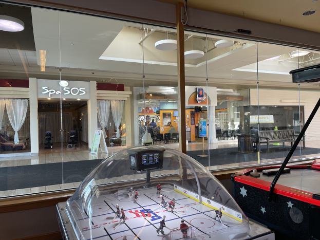Eine Eishalle in einem Einkaufszentrum, umgeben von Glaswänden mit Menschen, Möbeln und Auslagen, die durch die Glaswände sichtbar sind, und einem Air-Hockey-Tisch auf der rechten Seite.